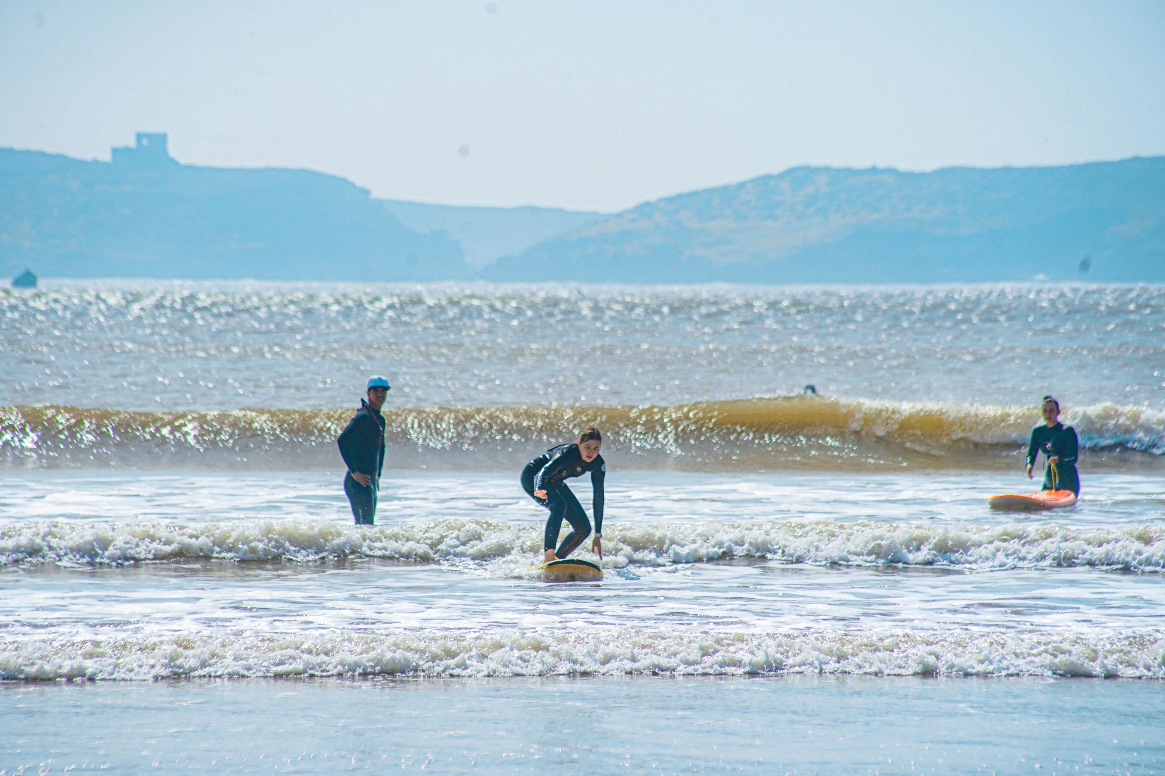 Élève surf Bluboarding essayant d'attraper une vague tandis que son moniteur observe et son amie regarde depuis la plage principale d'Essaouira.