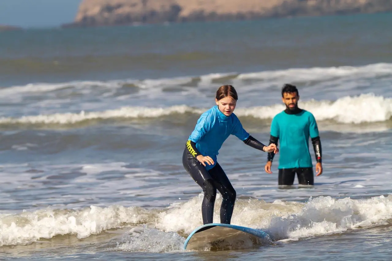 Jeune cliente Bluboarding attrapant une vague sur la plage principale d'Essaouira tandis que son moniteur de surf l'observe depuis l'arrière lors d'un cours privé.